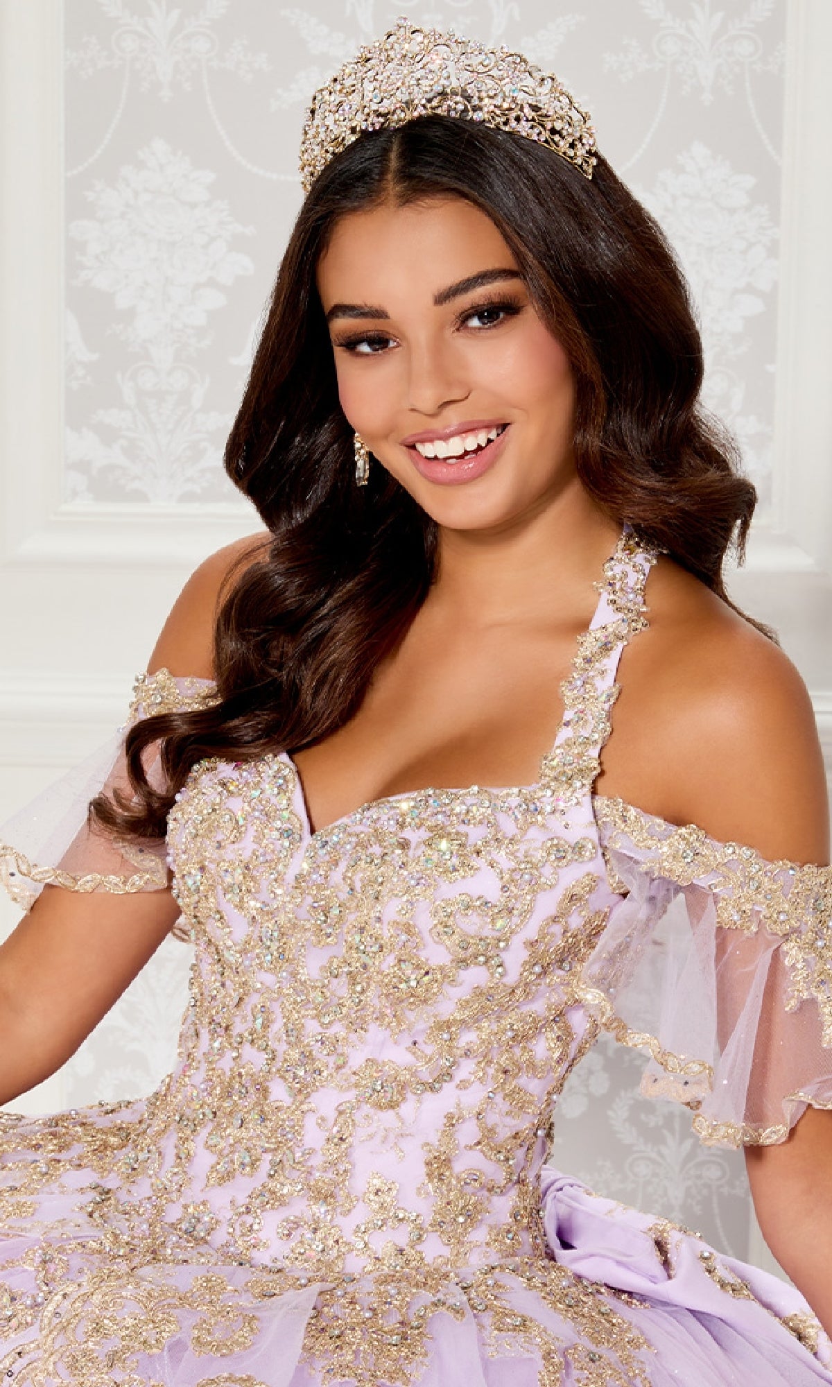 A woman with long wavy hair smiles at the camera, wearing a jeweled tiara and the ornate PR30118 Quinceanera Dress by Princesa—an off-the-shoulder lilac gown featuring gold embellishments and glitter embroidery.